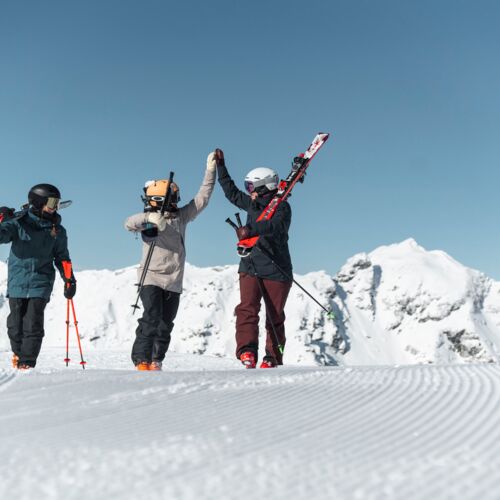 Drei Skifahrer mit geschulterten Ski vor verschneiten Berggipfeln klatschen ein